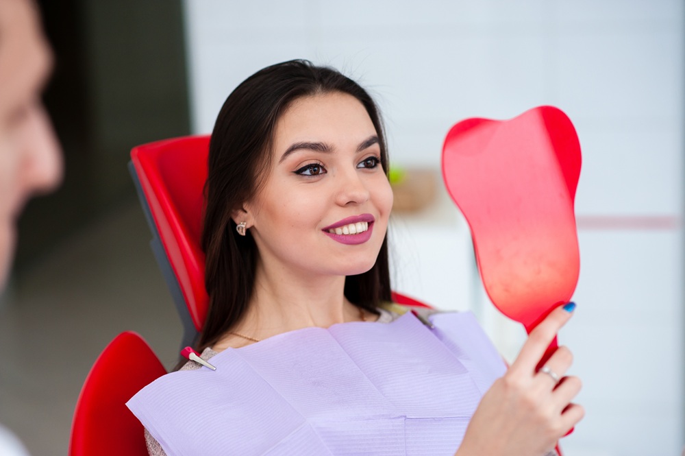 Patient Admiring Smile After Dental Treatment – Cosmetic Dentistry A patient smiles while looking in the mirror after a successful cosmetic dental procedure, pleased with her new bright and even smile. – Cosmetic Dentistry