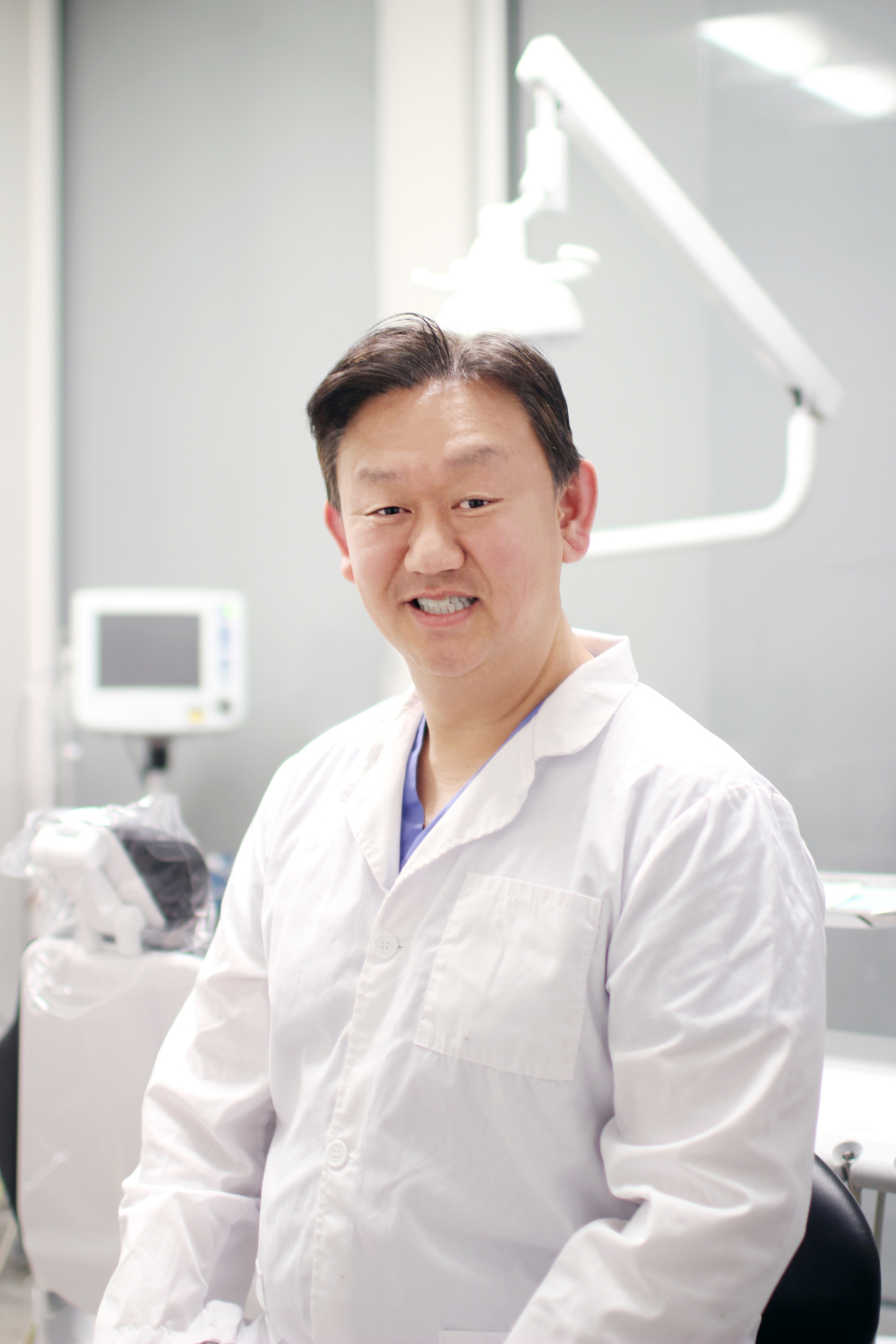 Dr. Chris Ko, DDS, smiling while wearing a white coat and seated in a modern dental operatory with equipment visible in the background – Dentist in Redmond WA