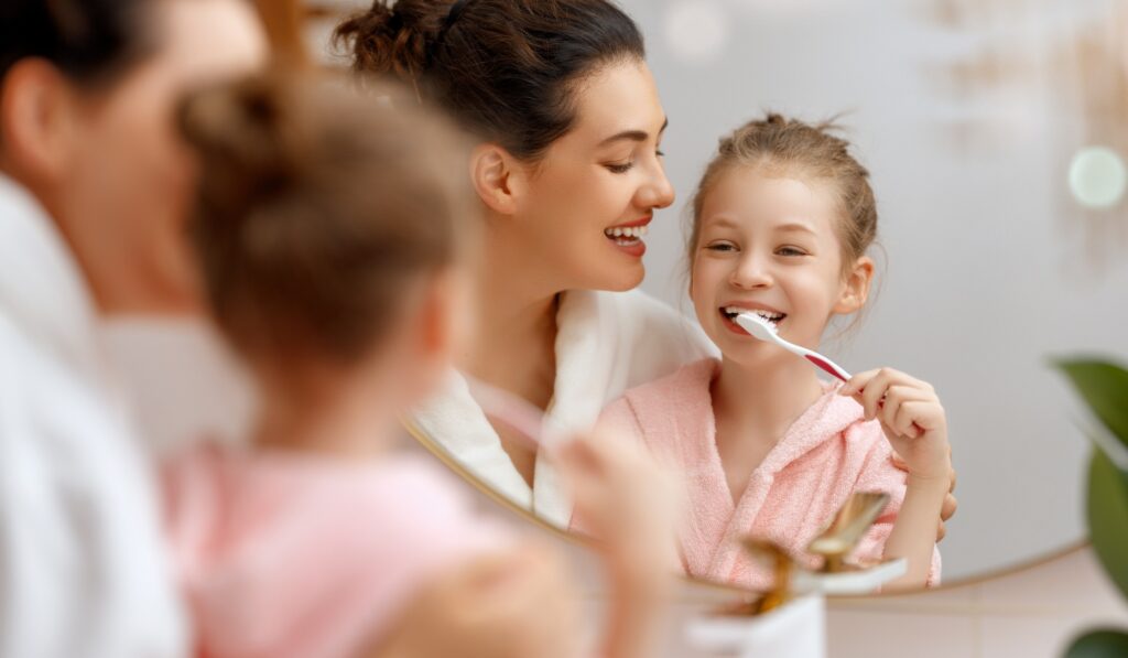 Mother and daughter brushing teeth together in bathroom, smiling and wearing bathrobes - Dentist in Redmond WA A mother and daughter, both smiling in bathrobes, brush their teeth together in a warmly lit bathroom, sharing a joyful, intimate moment. - Dentist in Redmond WA