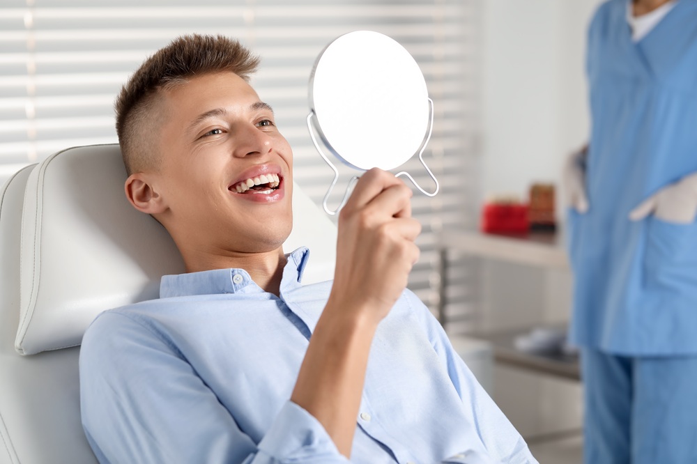 A young man smiles while looking into a mirror after his dental restoration, appreciating his improved smile with durable crowns or bridges. – Crowns & Bridges