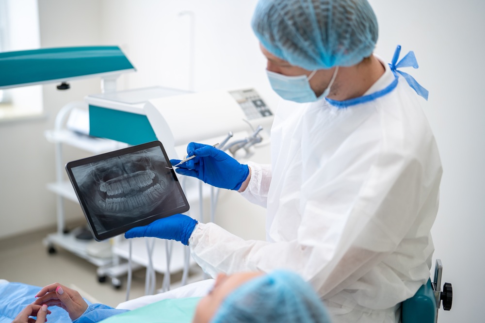 Dentist Reviewing Panoramic Scan on Tablet – Digital X-Ray A dentist in protective gear examines a patient’s panoramic dental X-ray on a tablet, using advanced digital imaging for accurate diagnosis and treatment planning. – Digital X-Ray