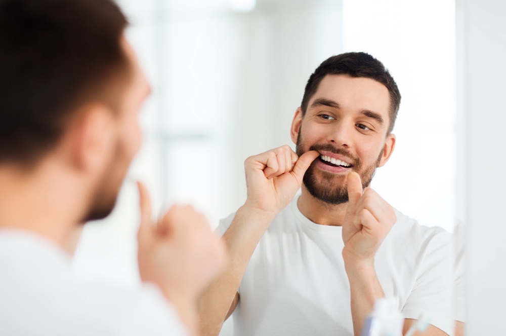 Man Flossing for Oral Hygiene Maintenance – Dental Cleaning in Seattle A man flosses his teeth in front of the mirror, maintaining his oral health and complementing the results of his recent dental cleaning. – Dental Cleaning in Seattle