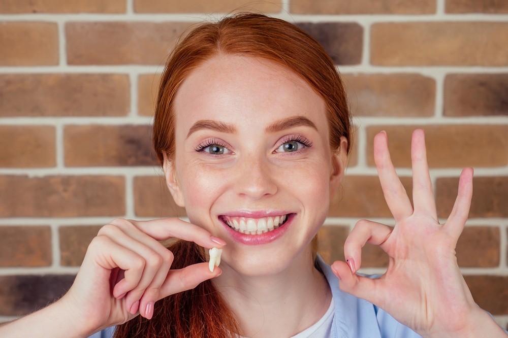Happy Patient After Tooth Extraction – Extractions A smiling woman holds a removed tooth and shows an OK gesture, representing a successful and painless dental extraction experience. – Extractions