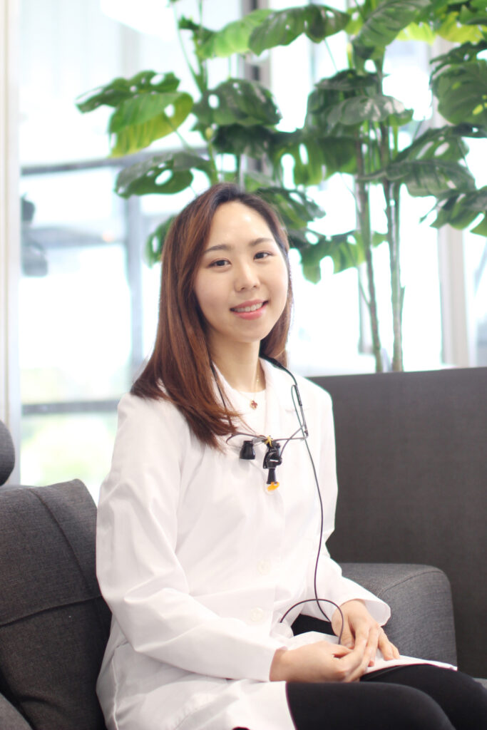 Dr. Su-Young Jane Choi, DDS, smiling while seated on a sofa in a bright dental office, wearing a white coat with dental loupes around her neck – Dentist in Redmond WA