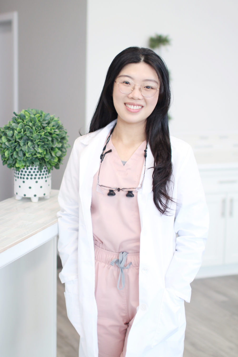 Dr. Yongxi Liang, DMD, smiling while standing in a bright dental office wearing a white coat over pink scrubs with dental loupes around her neck – Dentist in Redmond WA